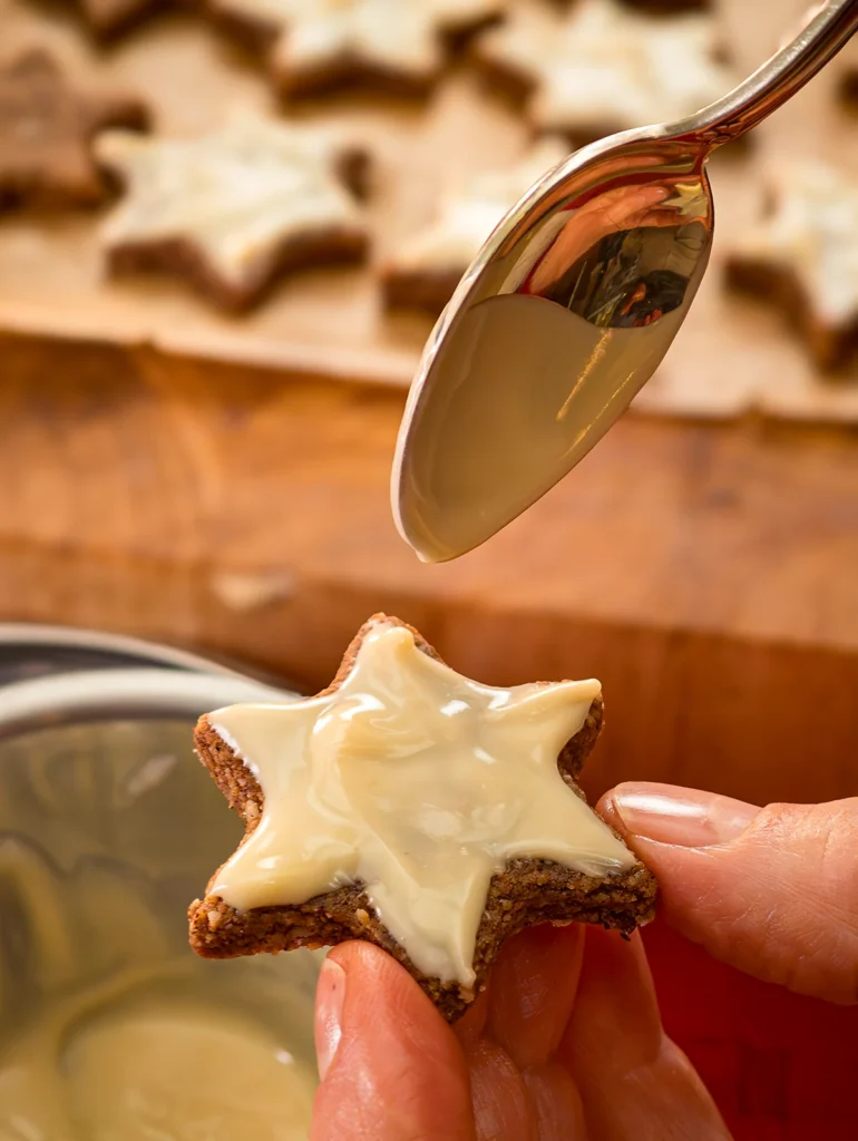 A hand holds a cookie and a spoon with melted chocolate is above it. The Cinnamon Star is ready glazed.