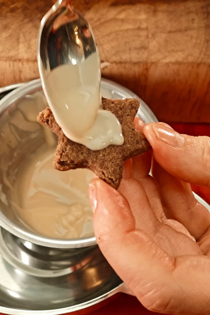 A hand holds a cookie and a spoon put a dollop of the melted chocolate coating in the middle of the Cinnamon Star to glaze it. Behind is the bain-marie with a bowl of melted white vegan chocolate.