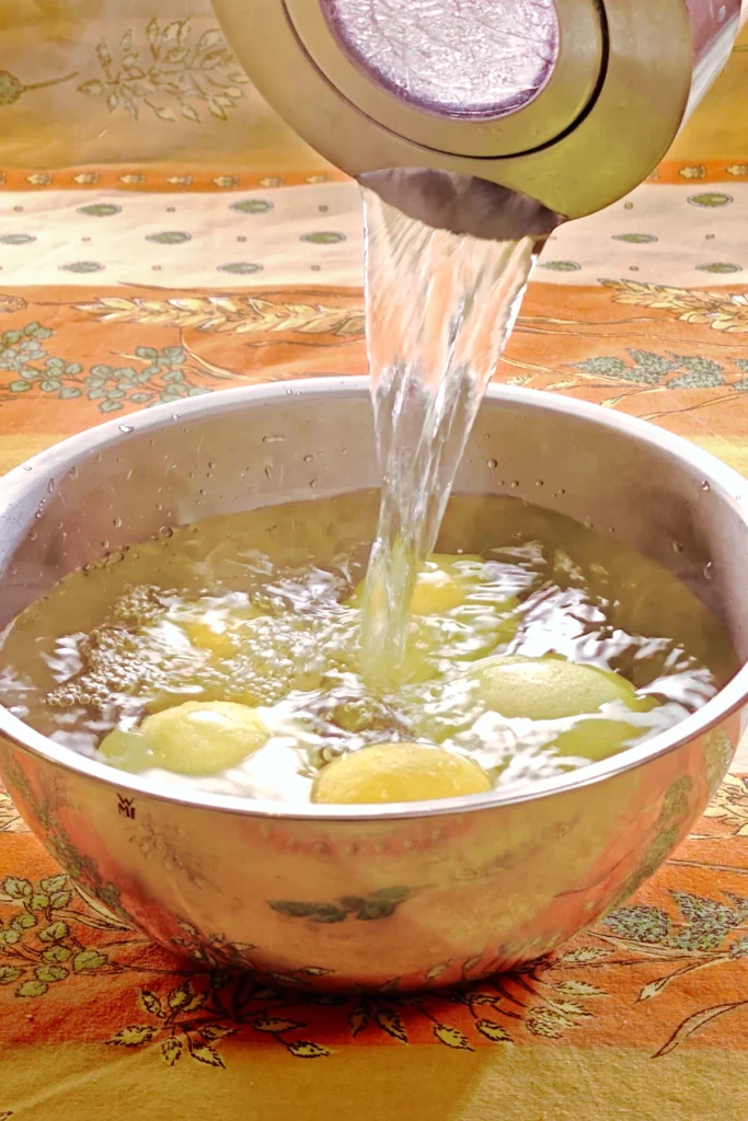 Lemons in a steel bowl are doused with hot water from a kettle. The bowl stands on a patterned orange yellow tablecloth.