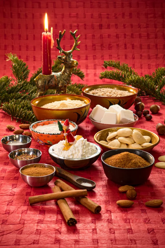 Festive baking ingredients for Cinnamon Star Cookies arranged on a red tablecloth, including ground almonds, ground hazelnuts, flour, corn starch, Ceylon cinnamon, coconut fat, coconut sugar, salt, flaxseed, baking powder, and vegan white chocolate. A holiday-themed setup with a candle, evergreen branches, and decorative elements.