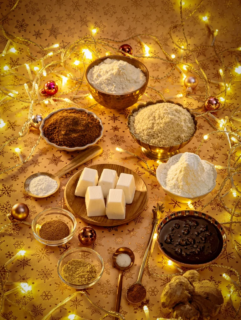 On a light background with festive gold star motifs, there are many bowls of ingredients for the gingerbread cookies. They contain corn starch, coconut sugar, flour, ground almonds, sugar-root molasses, and spices such as ground ginger and ground nutmeg. In the center is a wooden plate with pieces of coconut fat. On three spoons are cinnamon, salt and baking powder. In the foreground is a ginger root. All around is a string of lights and a few small Christmas tree baubles are scattered around.