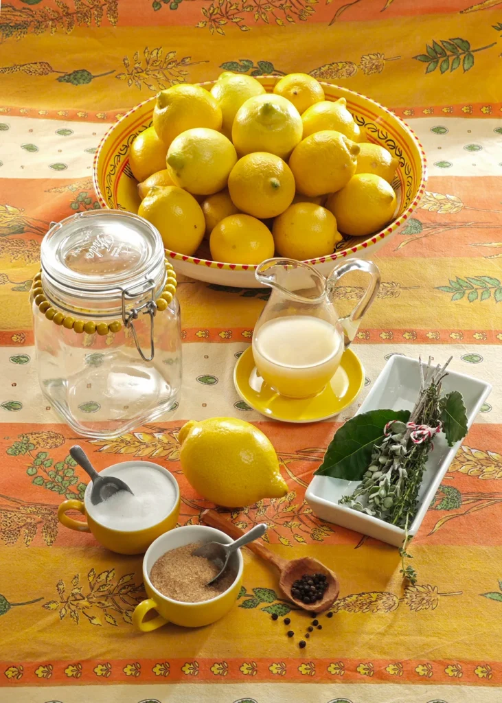 Bright yellow lemons in a decorative bowl, surrounded by ingredients for preserved lemons, including salt, sugar, lemon juice, black peppercorns, and a bundle of fresh herbs, displayed on a vibrant orange tablecloth with a clear preserving jar ready for use.