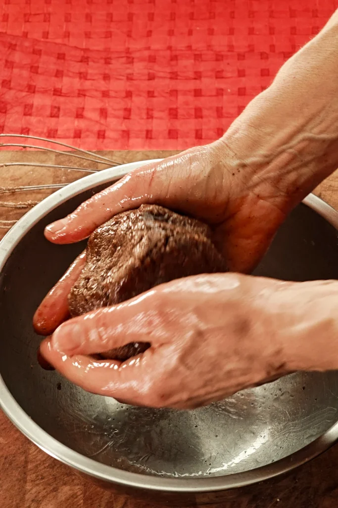 Two hands hold kneading the greasy dough into a ball. The background is a red tablecloth.