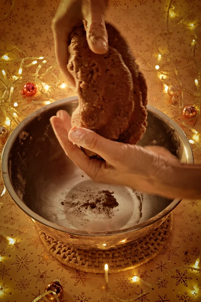 Two hands knead the gingerbread cookie dough over a stainless steel bowl. A string of lights shines in the background and a few small Christmas tree baubles are scattered around. The light background has a festive golden star motif.