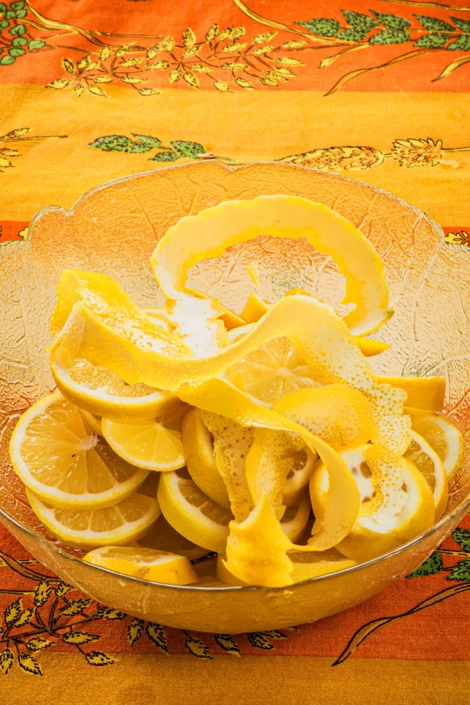 On an patterned orange yellow tablecloth is a glass bowl with lemon slices and lemon peel.