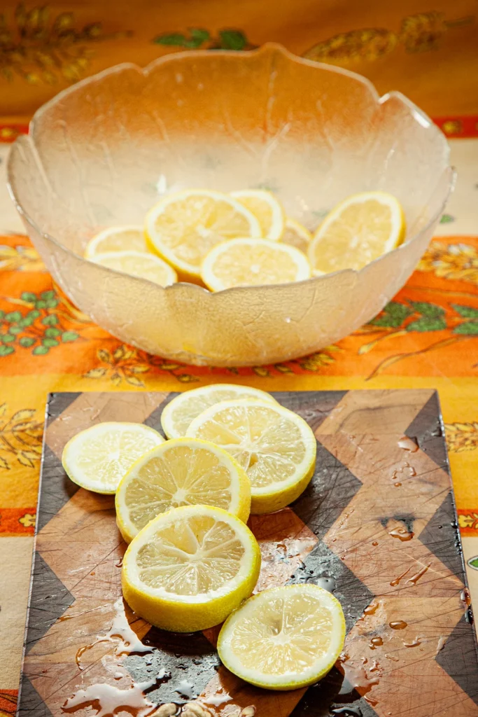 On an patterned orange yellow tablecloth lies a wooden kitchen board with lemon slices. Behind the kitchen board is a glass bowl with lemon slices.