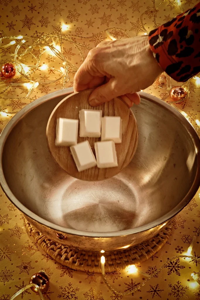 A hand add blocks of coconut fat into a stainless steel bowl. A string of lights shines in the background, and a few small Christmas tree baubles are scattered about. The light background has a festive gold star motif.