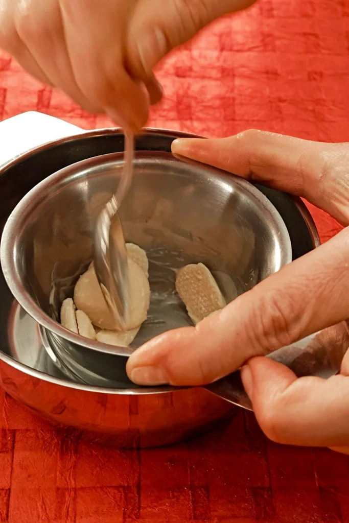 Two hands hold a bowl with vegan white chocolate coating, that stands in a hot water bain-marie. One hand holds a spoon, stirring in the melting chips. The water bowl stands on a red tablecloth.