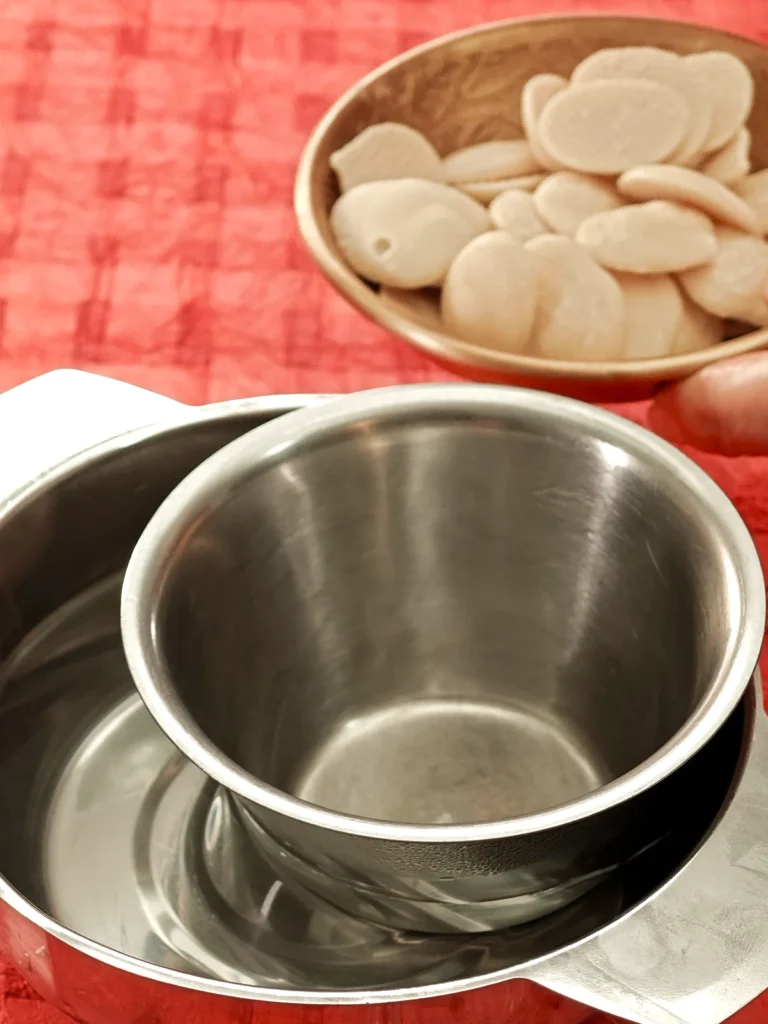 One hand holds a bowl of vegan white chocolate coating, ready to melt them it in a prepared water bain-marie. The water bowl stands on a red tablecloth.