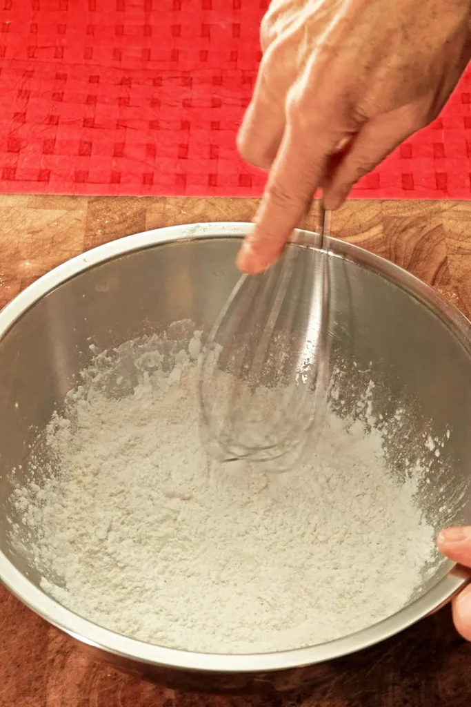 A hand is holding a whisk to mix the baking powder with the flour and starch in a stainless steel bowl. In the background you can see a red tablecloth.