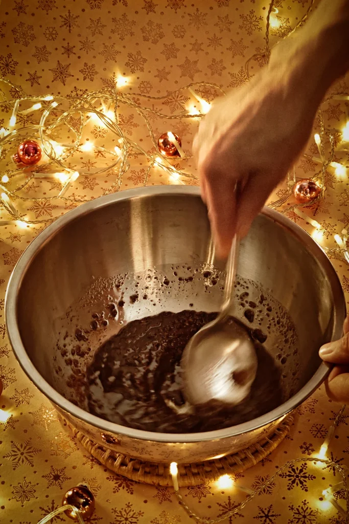 A hand with a silver spoon stirs coconut sugar and melted coconut oil in a stainless steel bowl. A string of lights shines in the background, and a few small Christmas tree baubles are scattered about. The light background has a festive gold star motif.