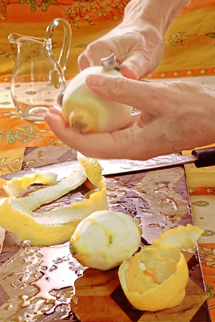 On an orange tablecloth lies a wooden kitchen board with lemon peel, a peeled lemon and a large knife. Behind it is an empty glass jug. Two hands are holding a lemon that has almost been peeled with the help of a potato peeling knife.