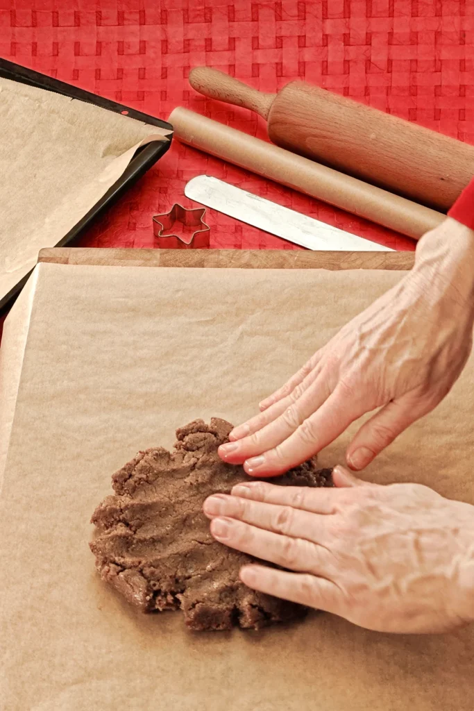 Two hands put the dough on baking paper. In the background you can see a red tablecloth with a baking tray that is also lined with baking paper, a rolling pin, a spatula and a star cookie cutter.