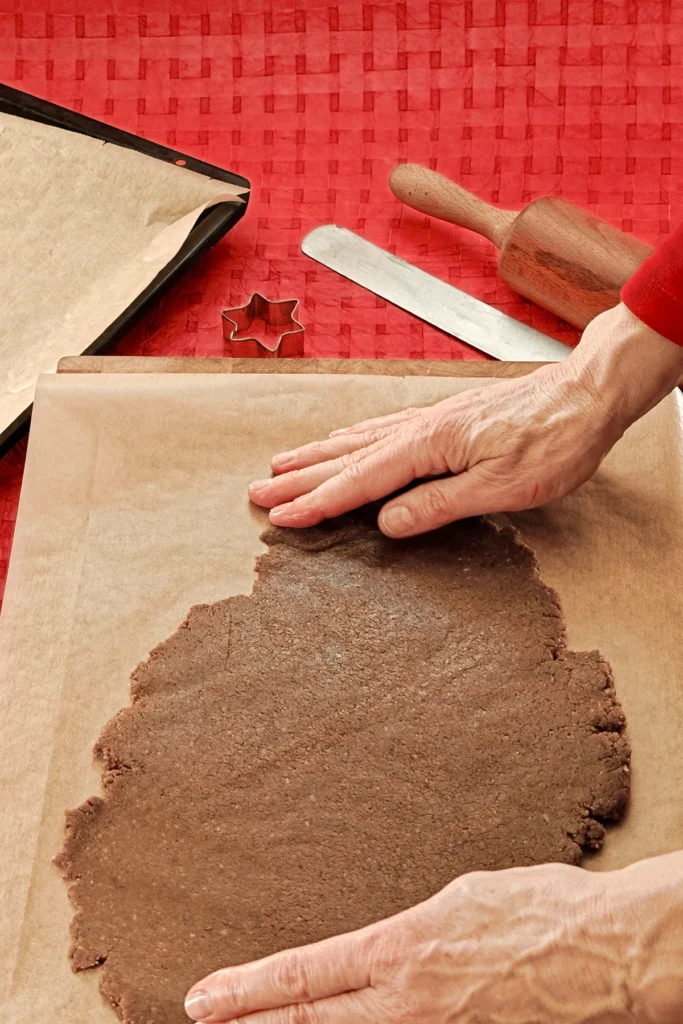 Two hands check the rolled out dough lying on baking paper. In the background you can see a red tablecloth with a baking tray that is also lined with baking paper, a rolling pin, a spatula and a star cookie cutter.