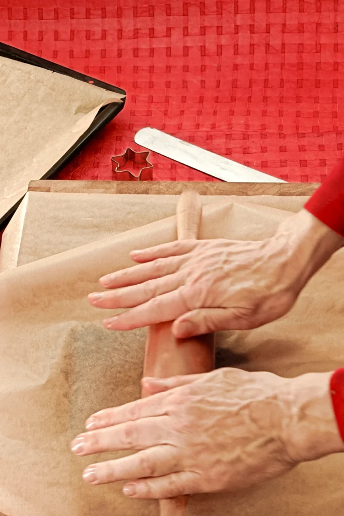 Two hands hold a rolling pin and roll the dough, which is placed between two baking papers. In the background you can see a red tablecloth with a baking tray that is also lined with baking paper, a spatula and a star cookie cutter.