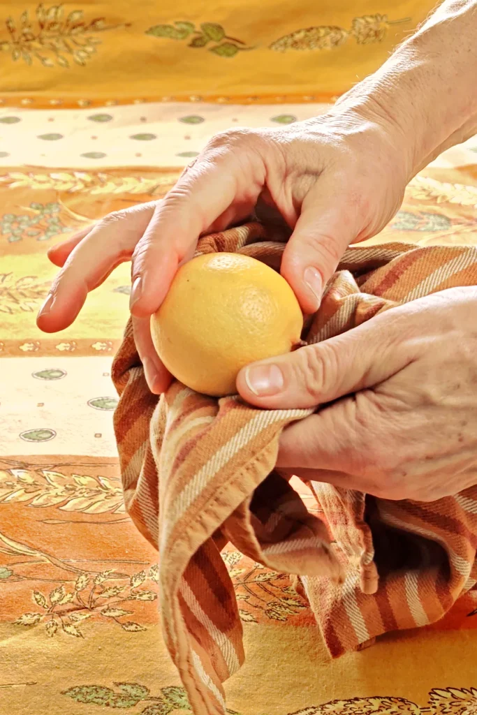 The lemon is dried by two hands with a striped kitchen towel. The background is a tablecloth in orange tones with wheat and oat straw motifs.