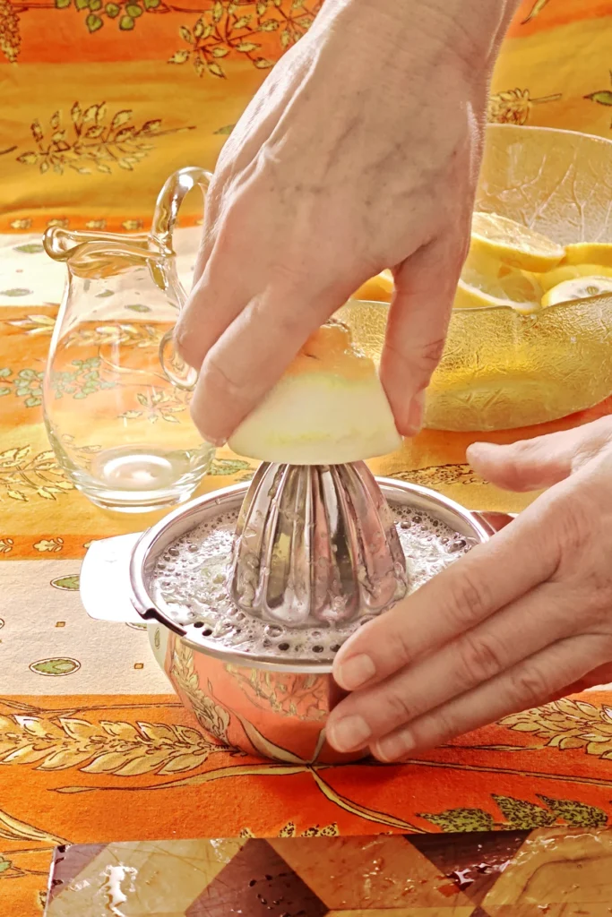 Lemon is squeezed on a citrus press. the press stands on a tablecloth in orange tones with wheat and oat straw motifs. Behind the press is a glass bowl with lemon slices.