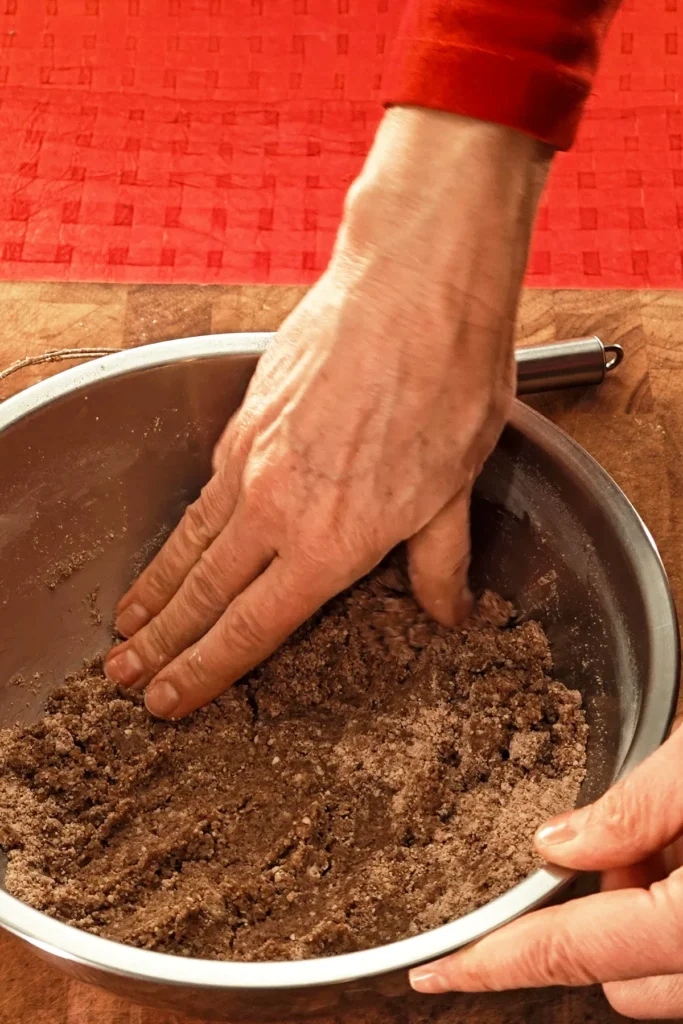 Two hands hold the stainless steel bowl and knead the dough. The background is a red tablecloth.