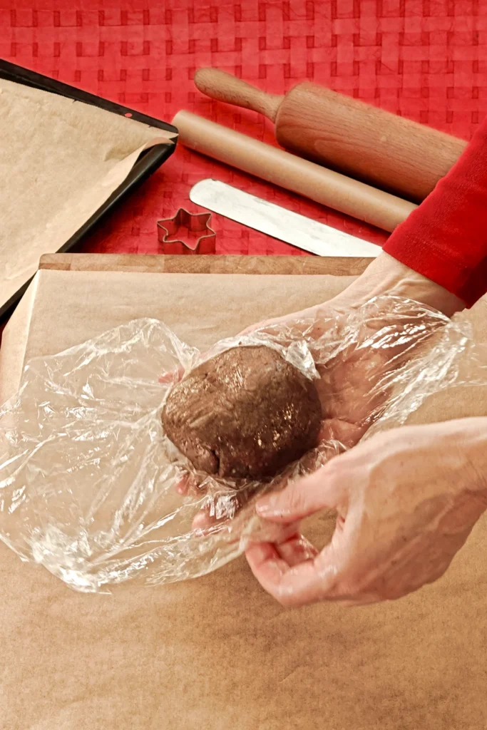 Two hands hold the ball of dough and unwrap it from the plastic wrap. A kitchen board with baking paper is placed underneath. In the background you can see a red tablecloth with a baking tray that is also lined with baking paper, a rolling pin, a spatula and a star cookie cutter.