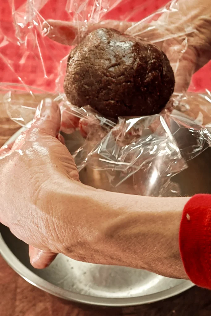 Two hands hold the ball of dough wrapped in plastic wrap. Underneath is the stainless steel bowl in which the dough was kneaded. The background is a red tablecloth.