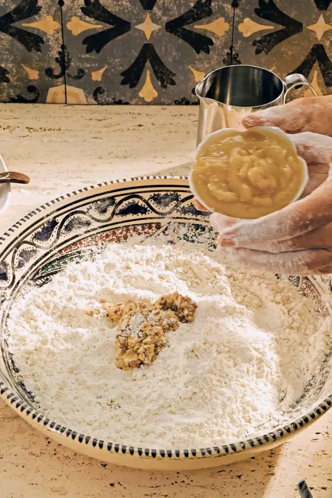 A large patterned ceramic bowl with a flour mixture and flaxseeds stands on a light-colored travertine kitchen table. Floured hands hold a white porcelain bowl with apple puree above it, preparing dough for vegan almond biscotti with orange and marzipan.