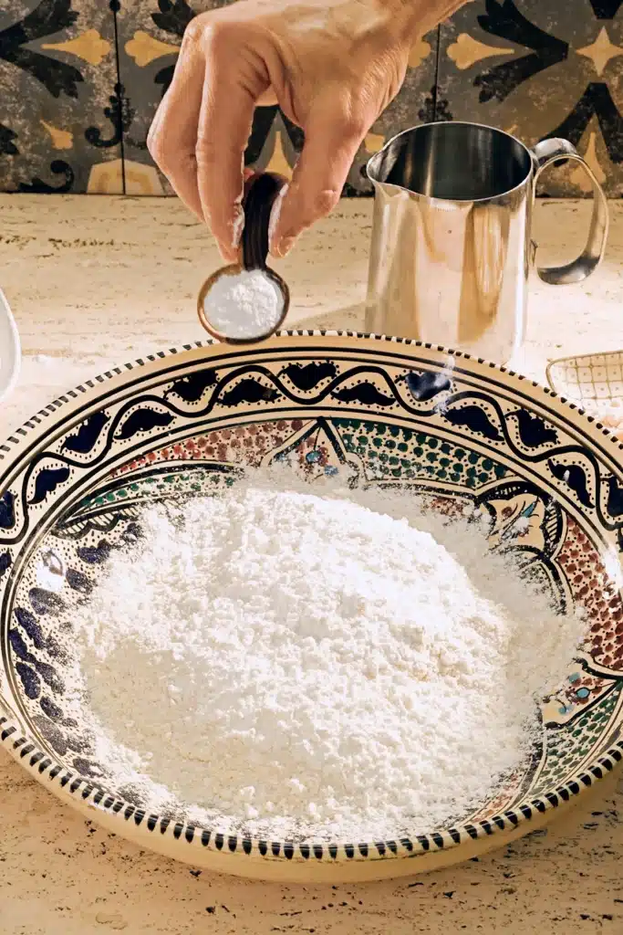 A large patterned ceramic bowl with a flour mixture stands on a light-coloured travertine kitchen table. A floured hand holds a wooden spoon with baking powder above it, preparing a dough for vegan almond biscotti with orange and marzipan.