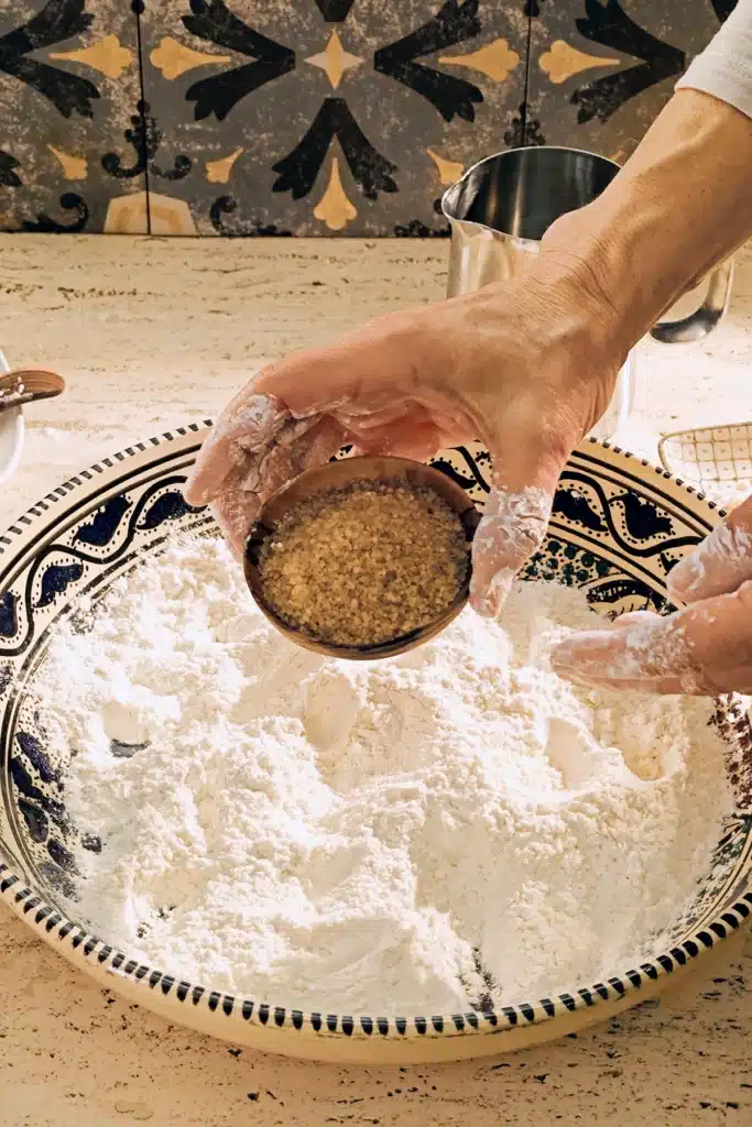 A large patterned ceramic bowl with a flour mixture stands on a light-coloured travertine kitchen table. A floured hand holds a wooden bowl with cane sugar over it, to make a dough for the vegan almond biscotti with orange and marzipan.