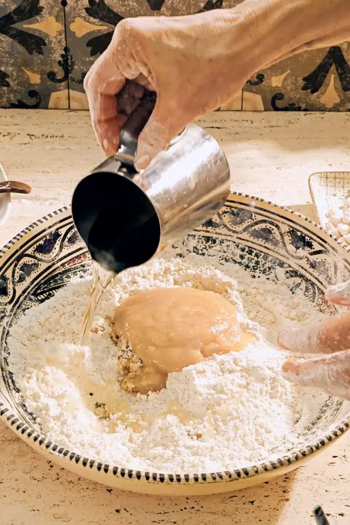 A large patterned ceramic bowl of flour and apple sauce stands on a light-coloured travertine kitchen table. A floured hand holds a stainless steel jug of coconut oil and pours it into the bowl.