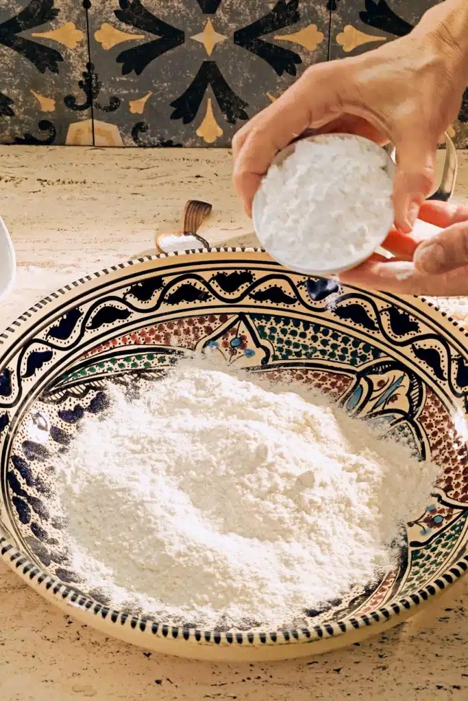 A large patterned ceramic bowl of flour stands on a light-coloured travertine kitchen table. Two hands hold a white bowl of cornflour over the bowl.