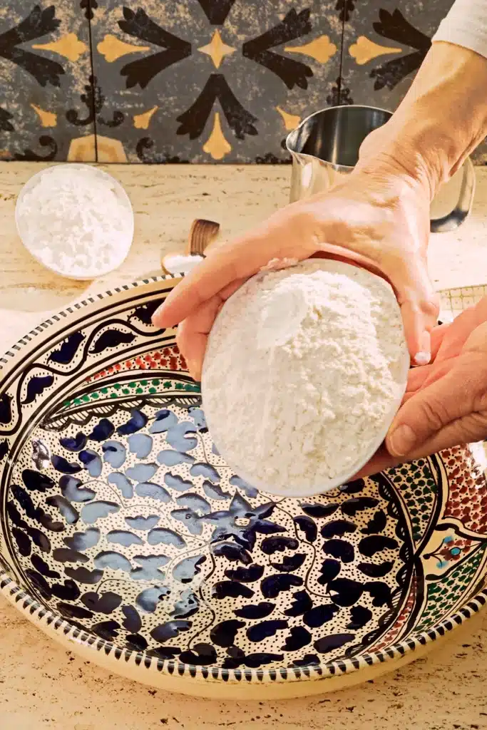 A large patterned ceramic bowl stands on a light-coloured travertine kitchen table. Two hands hold a white bowl of flour over the bowl.