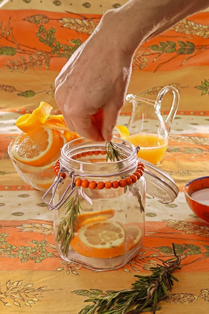 A hand adds rosmarin twigs to the preserving jar. Behind the jar is a glass bowl with orange slices, a jug with orange juice and a bowl with the salt and sugar mixture, all set on an orange tablecloth with wheat and oat straw motifs.