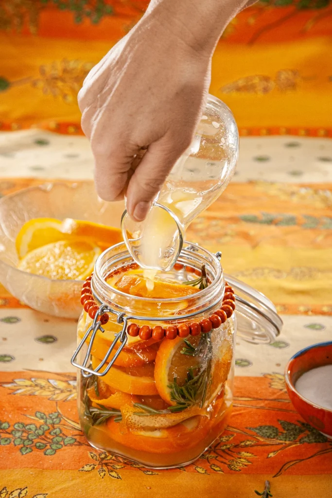 A hand is filling the preserving jar with orange juice from a glass jug. Behind is a glass bowl with orange slices, and an orange bowl with the salt and sugar mixture, all set on an orange tablecloth with wheat and oat straw motifs.