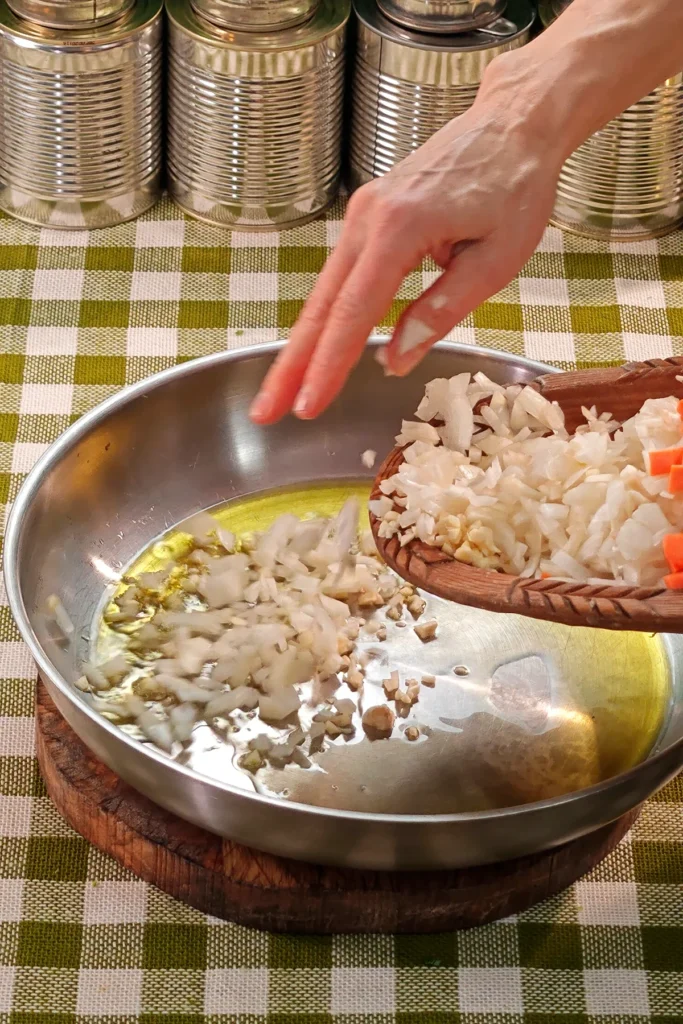 Add onions to the pan. The pan is placed on a round wooden board on a green chequered tablecloth. In the background is a row of tins.