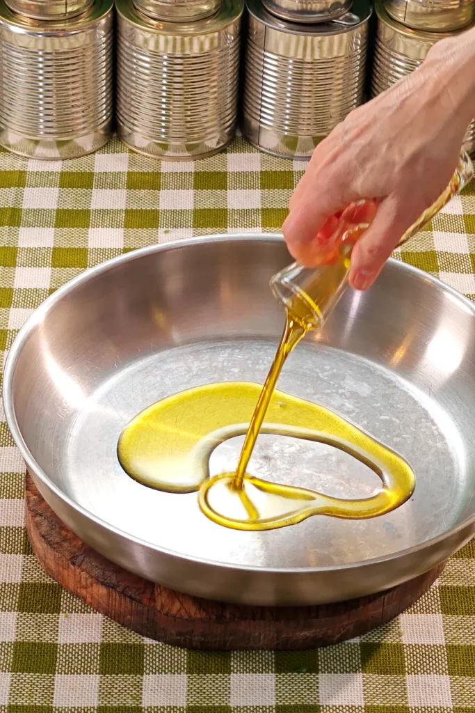 One hand holds a glass jug with olive oil and pures it into a pan. The pan is placed on a round wooden board on a green checkered tablecloth. A row of tins stands in the background.