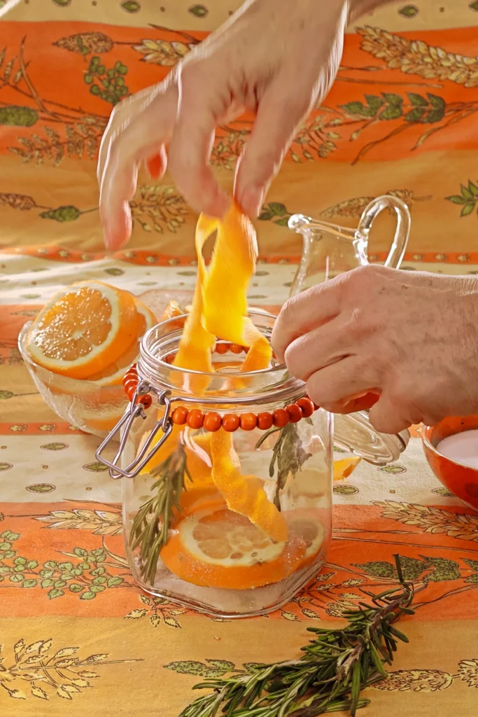 A hand is filling the preserving jar with a long orange peel. Behind the jar is a glass bowl with orange slices, a jug and a bowl with the salt and sugar mixture, all set on an orange tablecloth with wheat and oat straw motifs.