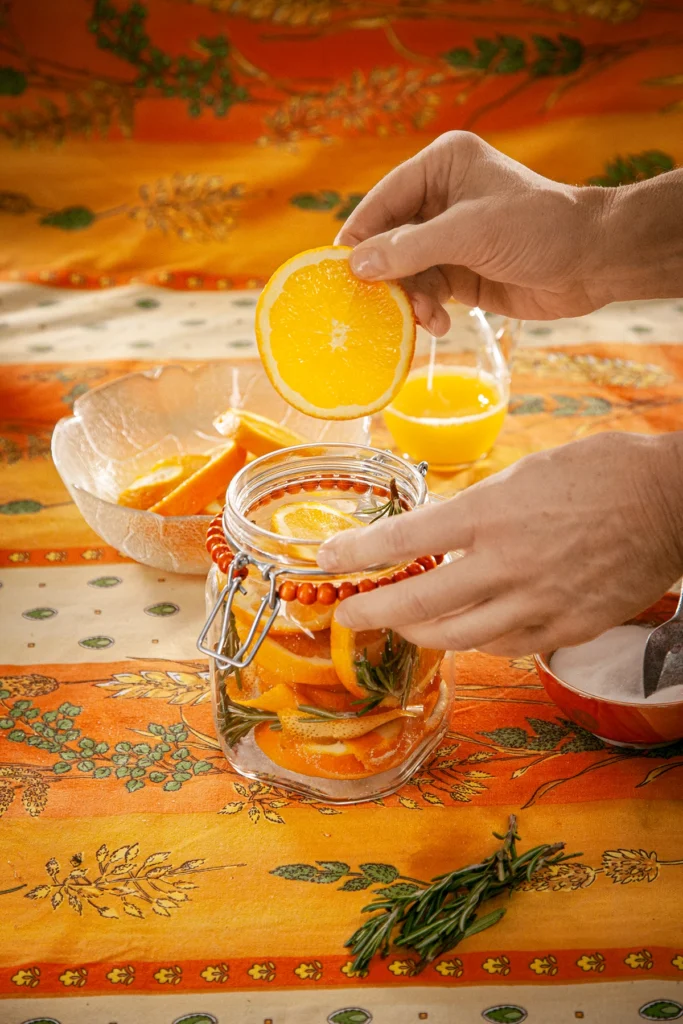 A hand is filling the preserving jar with orange slices. In front a some rosmarin twigs. Behind the jar is a glass bowl with orange slices, a jug with orange juice and a orange bowl with the salt and sugar mixture, all set on an orange tablecloth with wheat and oat straw motifs.