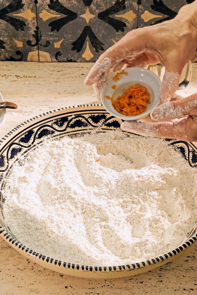 A large patterned ceramic bowl with a flour mixture stands on a light-coloured travertine kitchen table. Two floured hands will put grated orange peel from a small white bowl into the bowl.