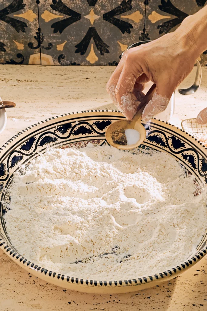 A large patterned ceramic bowl with a flour mixture stands on a light-coloured travertine kitchen table. A floured hand holds a wooden spoon with ground koriander and salt, to add it to the flour. On the wall in the background are grey patterned tiles.