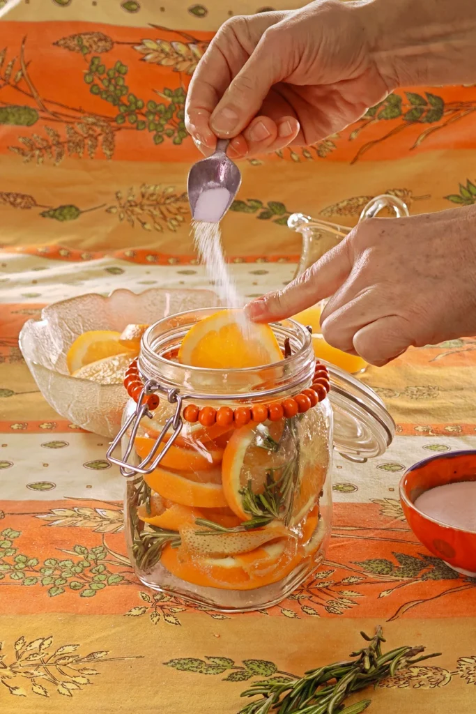 A hand adds the salt and sugar mixture from a spoon into the preserving jar. Behind the jar is a glass bowl with orange slices, a jug with orange juice and a bowl with the salt/sugar mixture - all set on an orange tablecloth with wheat and oat straw motifs.