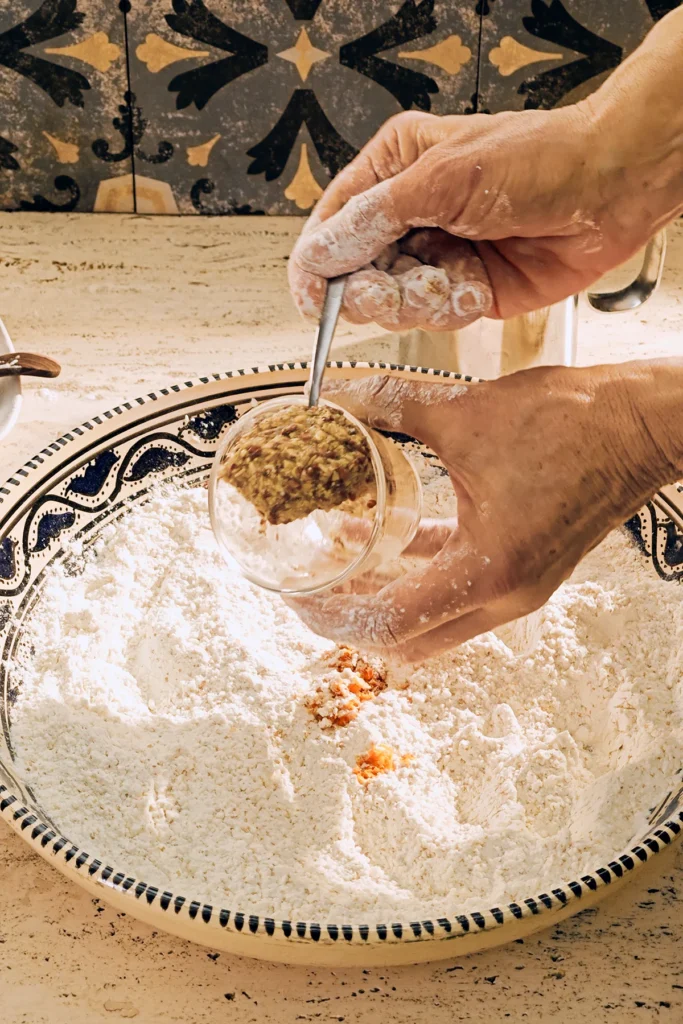 A large patterned ceramic bowl with a flour mixture stands on a light-coloured travertine kitchen table. Two floured hands add swollen flaxseed with a spoon from a glass.