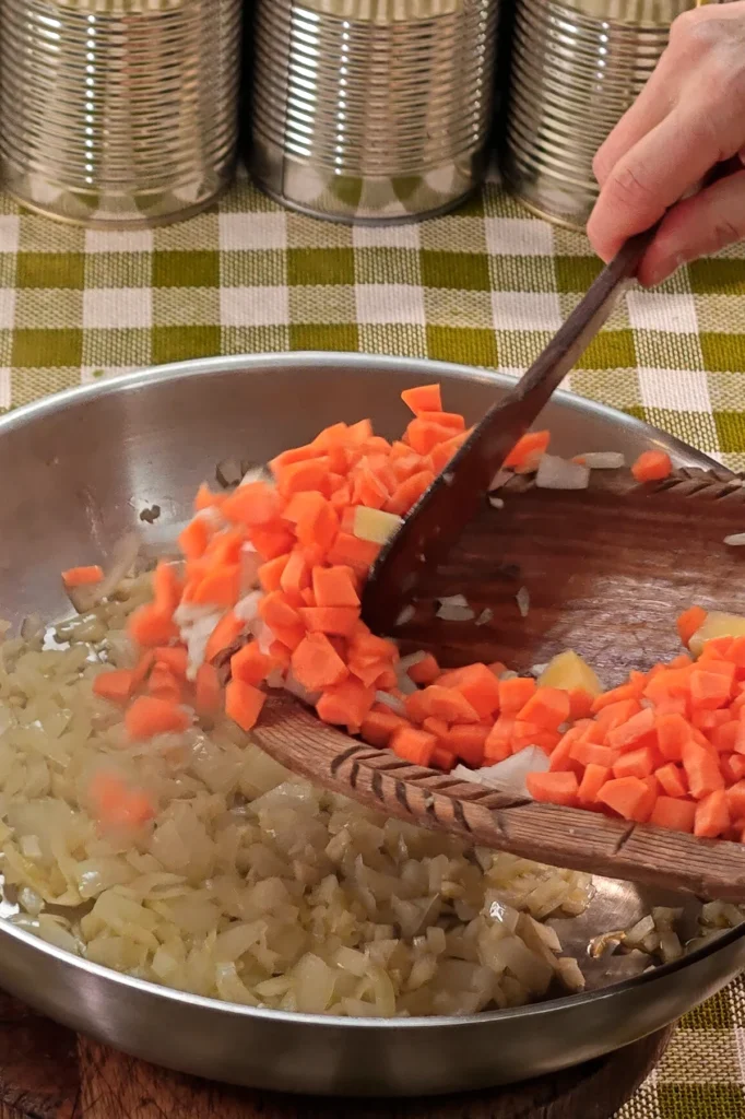 Add carotts to the pan. The pan is placed on a round wooden board on a green chequered tablecloth. In the background is a row of tins.