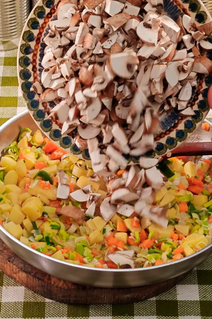 Add cremini mushrooms to the vegetable mixture. The pan is placed on a round wooden board on a green chequered tablecloth. In the background is a row of tins.