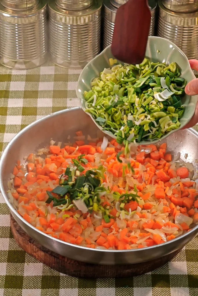 Add leek to the vegetable mixture. The pan is placed on a round wooden board on a green chequered tablecloth. In the background is a row of tins.