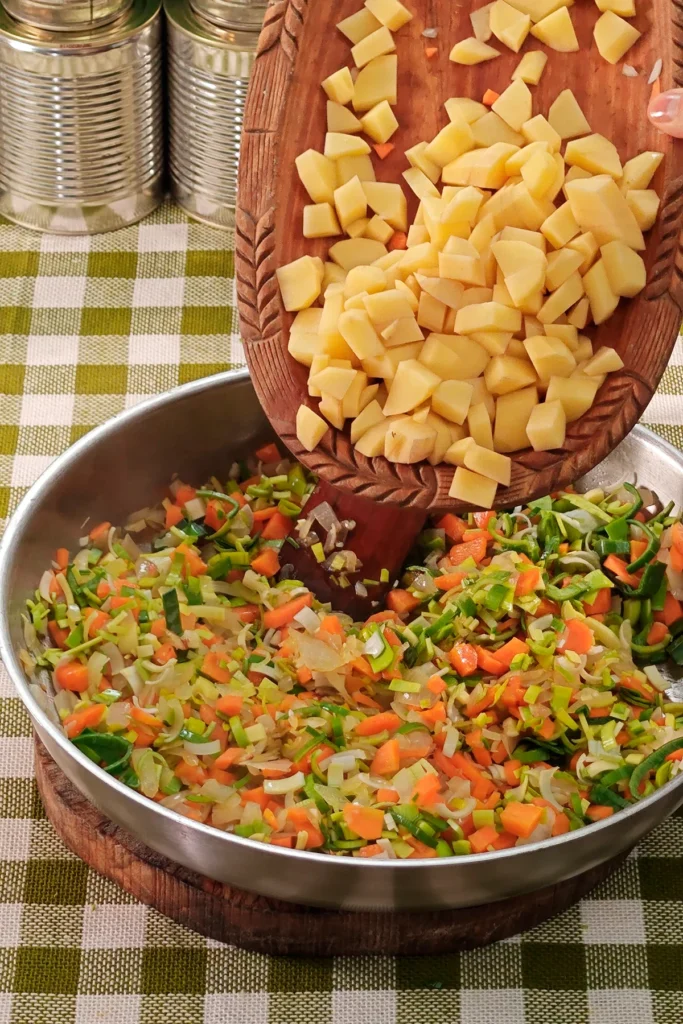 Add potatoes to the vegetable mixture. The pan is placed on a round wooden board on a green chequered tablecloth. In the background is a row of tins.