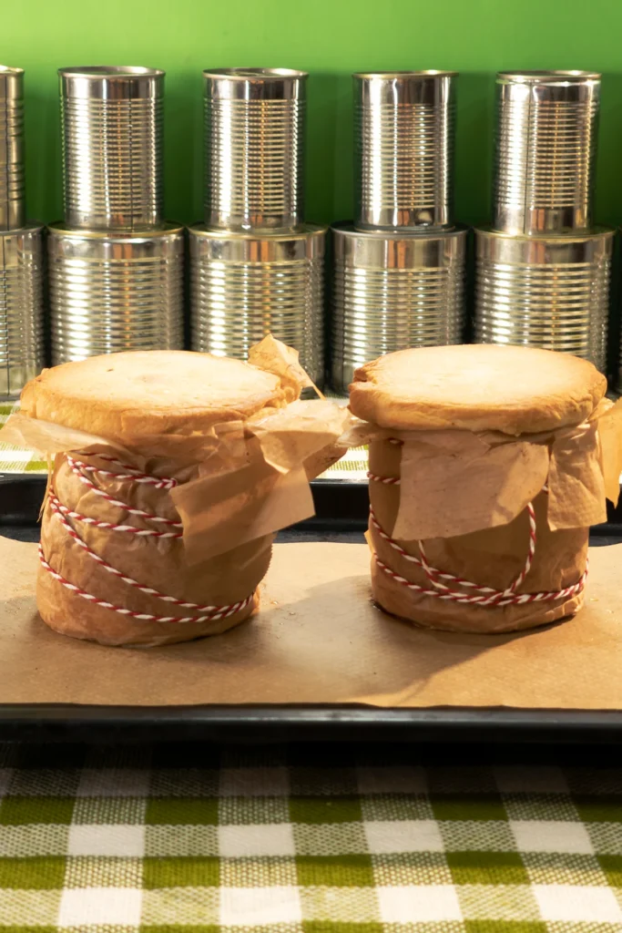 Golden-baked vegan jackfruit pie with a flaky crust and hearty filling. The two ready-baked savoury jackfruit pies are still wrapped in baking paper on the baking tray with baking paper. In the background is a row of tins.