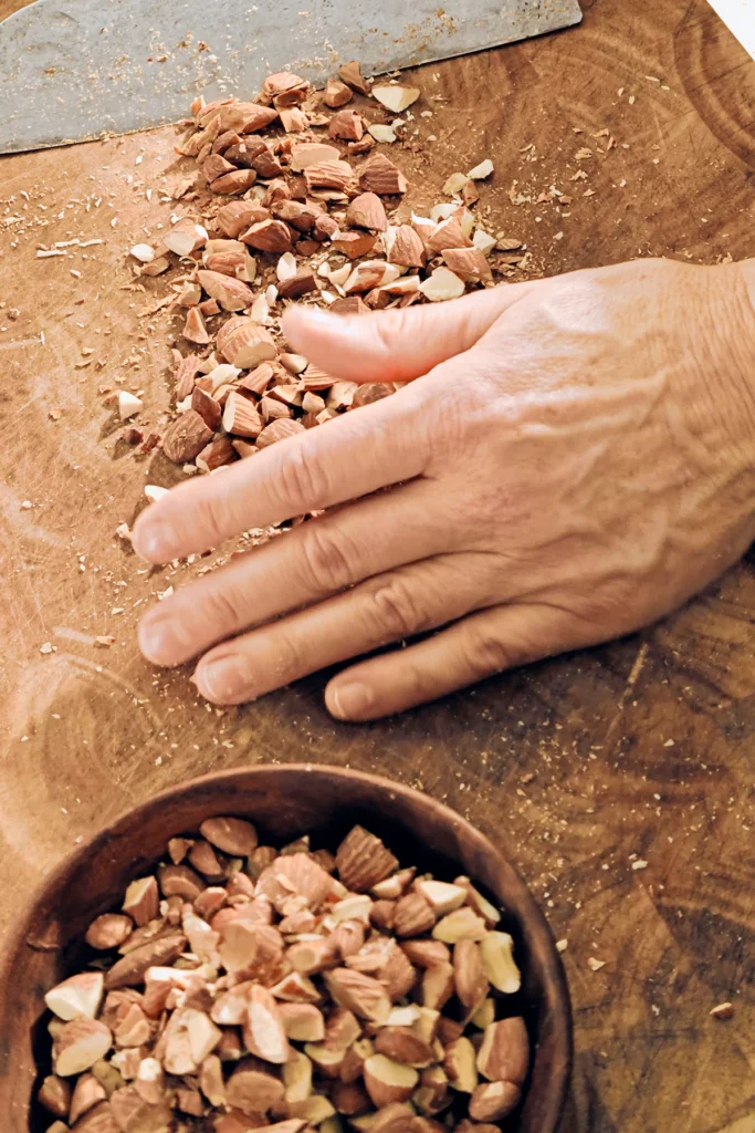 There are lots of roughly chopped almonds on a large wooden kitchen board. A knife blade and a hand push the pieces together. In front of them is a wooden bowl already filled with almond pieces.