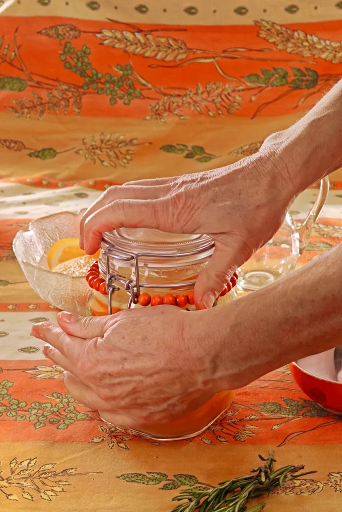 Close-up of a closed preserving jar with orange slices and herbs. The jar with the preserved oranges is standing on an orange and yellow patterned tablecloth. Two hands are in the process of closing the swing stopper on the lid. Behind is a glass bowl with left-over orange slices and an emty jug. In front is a bunch of rosmarin springs.