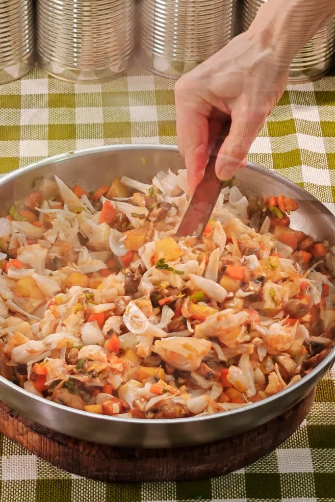 The savoury jackfruit and vegetable mixture is stirred and then steamed further. The pan is placed on a round wooden board on a green chequered tablecloth. In the background is a row of tins.