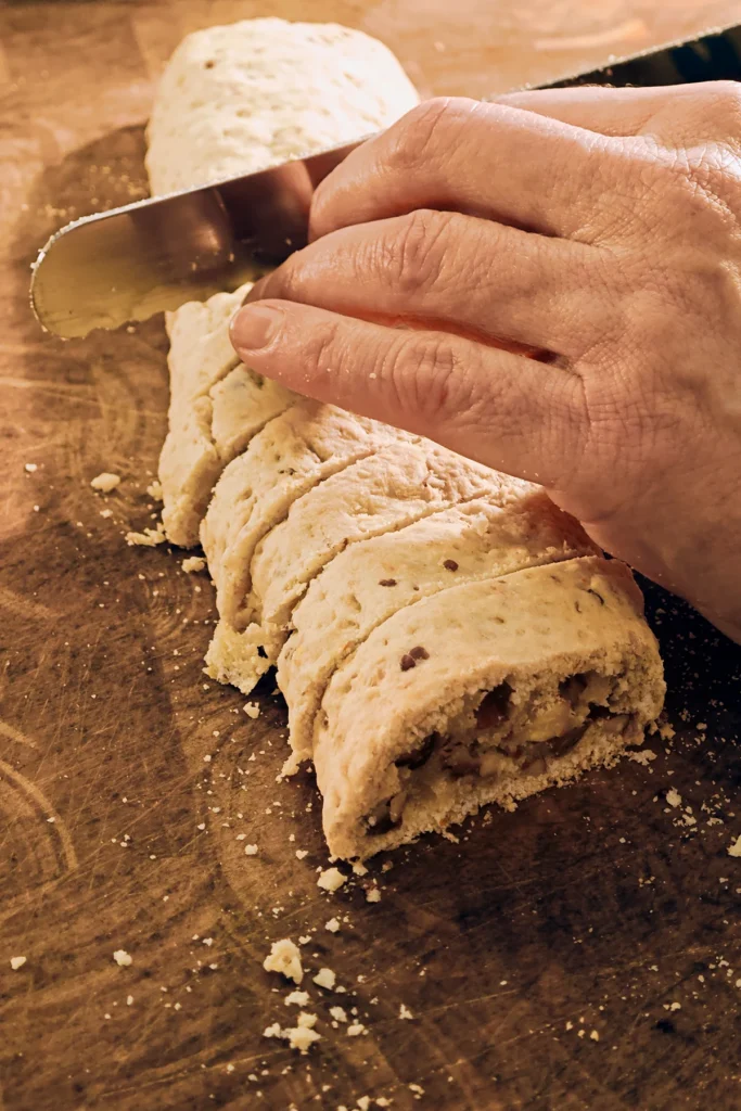 On a large wooden kitchen board lies a long roll of the prebaked vegan almond biscotti with orange and marzipan dough. Two hands are holding a large serragated knife, and cut the roll into slices.