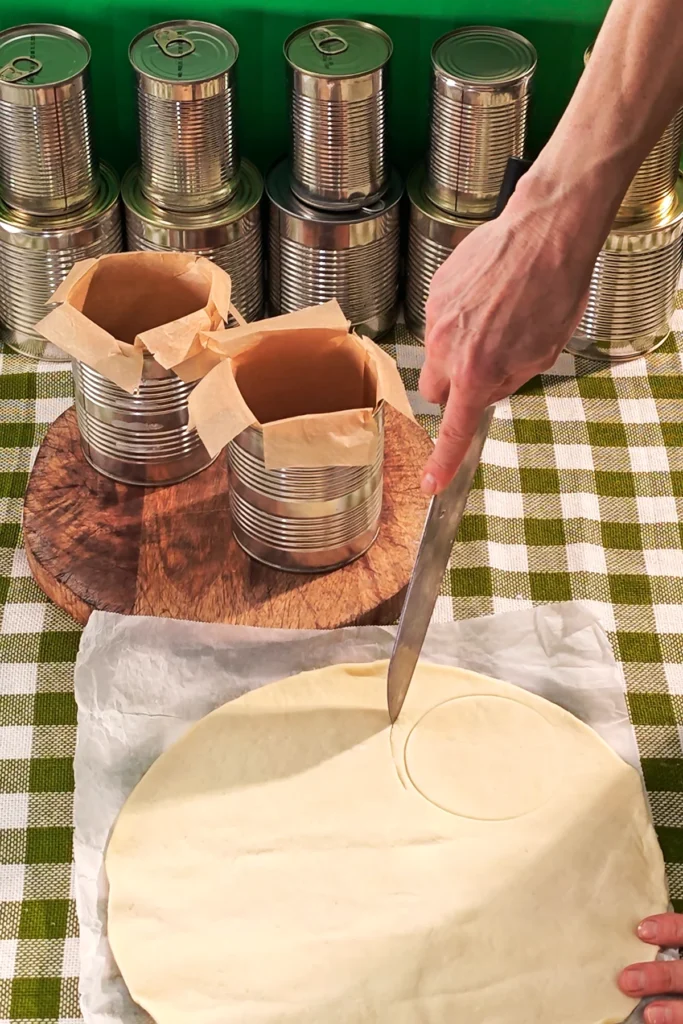 A roll of quiche dough is laid out on a green chequered tablecloth. A circle of dough is cut out around a circular punch with a large knife. Behind it is a round wooden kitchen board on which two tin cans lined with baking paper are placed. In the background is a row of tins.